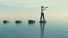 Young man on a large stone in the middle of the sea looking through binoculars